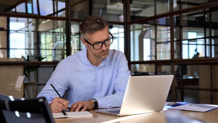 A well-dressed male employee writes in a notebook while looking at his computer