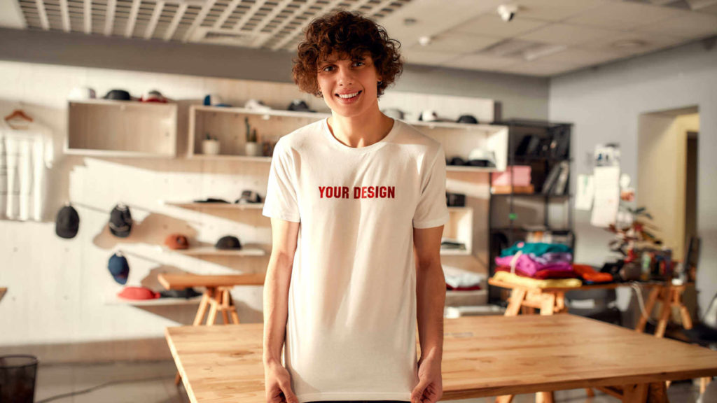 A young man smiles at the camera while wearing a white t-shirt reading “your design”