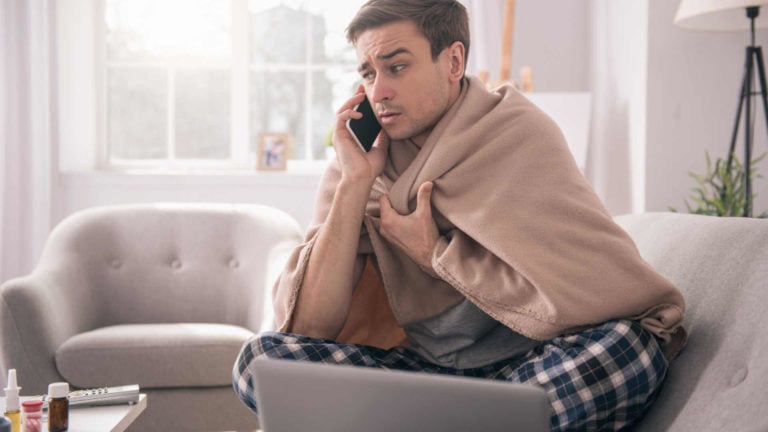 A sick employee sits on the couch with his computer and calls in sick to work