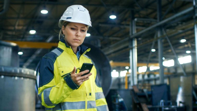A female factory worker wearing a hard hat and high-vis jacket looks at her phone