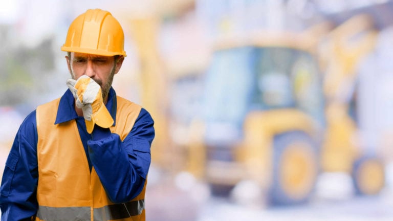 A sick construction worker coughs in his hand with a digger in the background