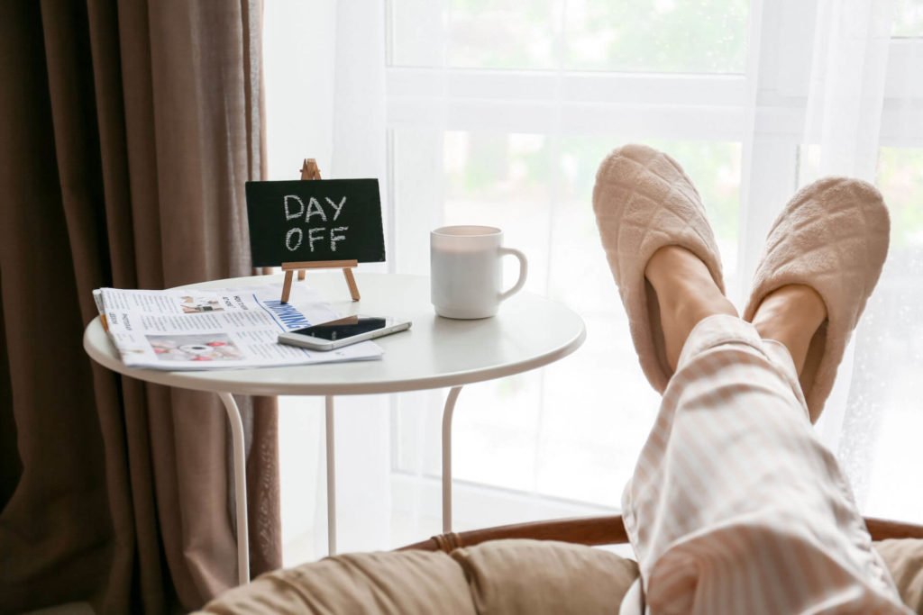 A woman wearing pyjamas sits at home next to a table with a sign reading “day off.”