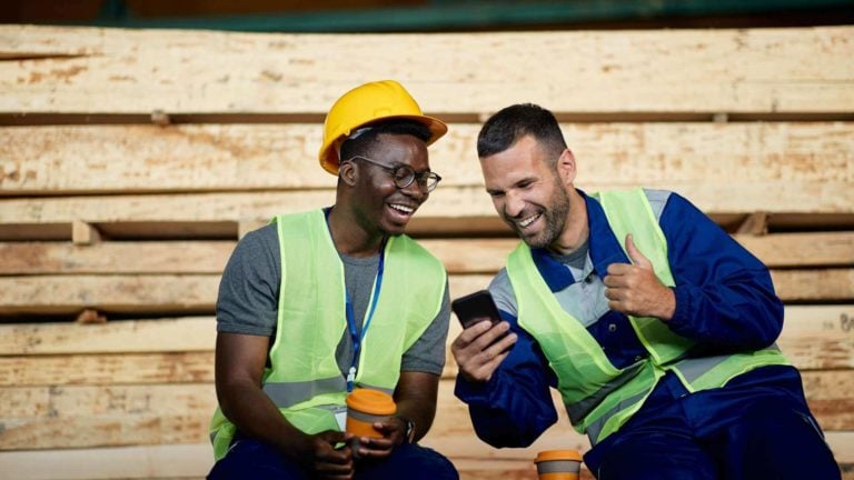 Two construction workers site near a pile of wood and laugh with a phone in their hand.