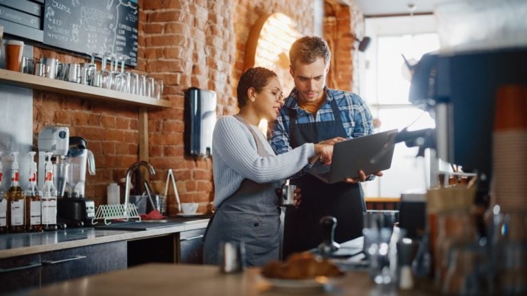 Two coffee sho workers stand over a laptop in a cafe