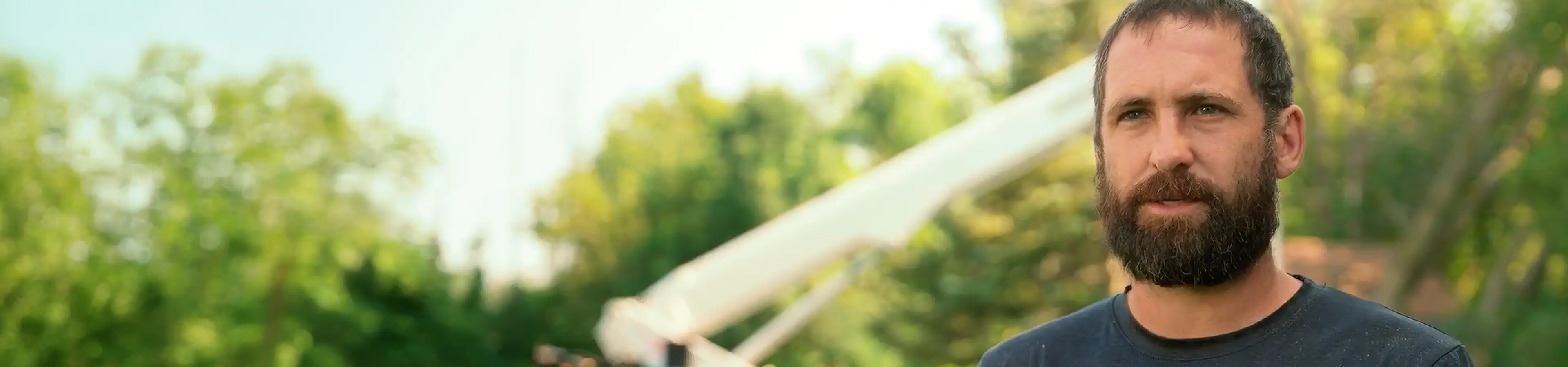 Bearded man outdoors with trees and an aerial lift in the background.