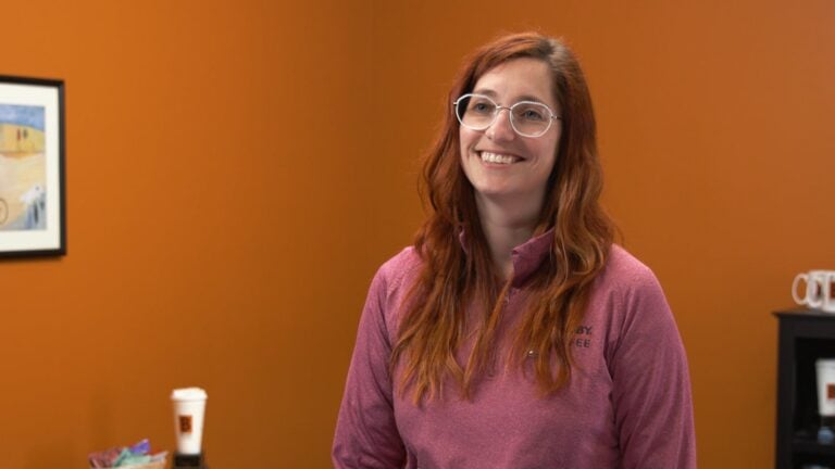 Smiling red-haired woman wearing glasses and a pink quarter-zip shirt against an orange wall.